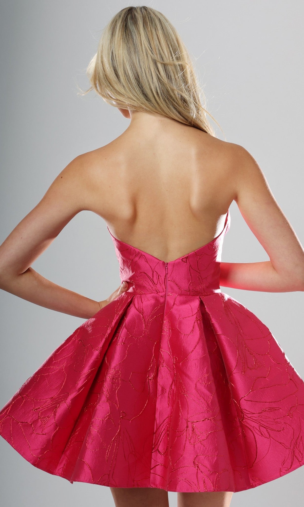 A woman with long blonde hair is shown from the back against a plain background, wearing the Jovani 52120 Short Homecoming Dress—a strapless, bright pink fit-and-flare dress featuring a floral pattern.